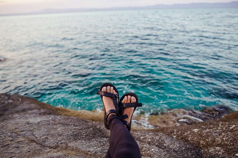 picture of woman beside a water sitting on a cliff, wearing orthotic sandals 